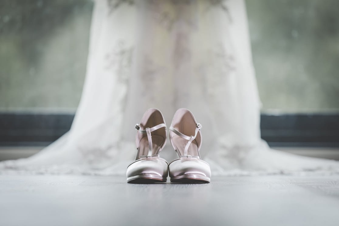 bride's shoes on floor with dress hanging up behind